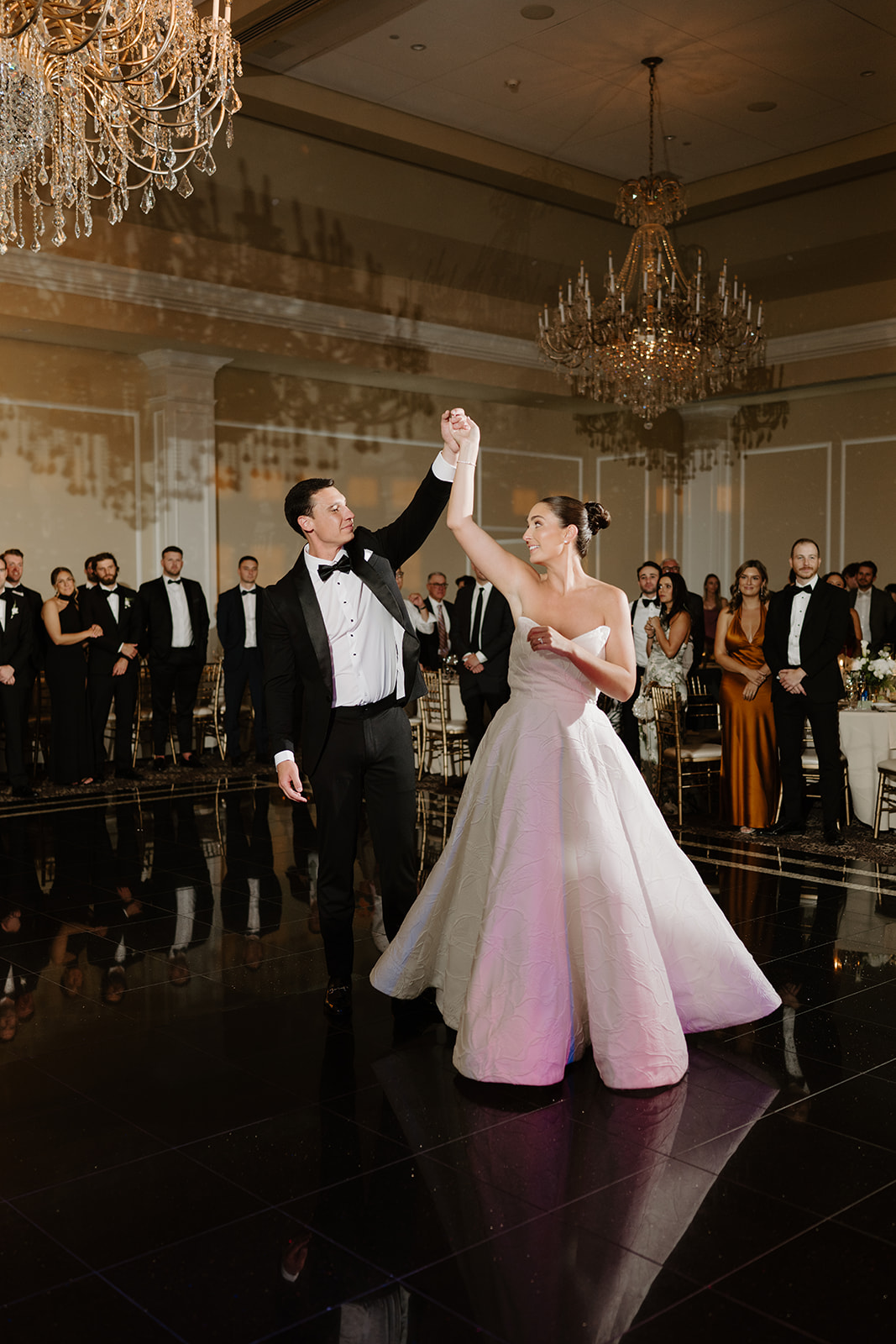 Bride and groom sharing their first dance on a glossy black floor under grand crystal chandeliers.